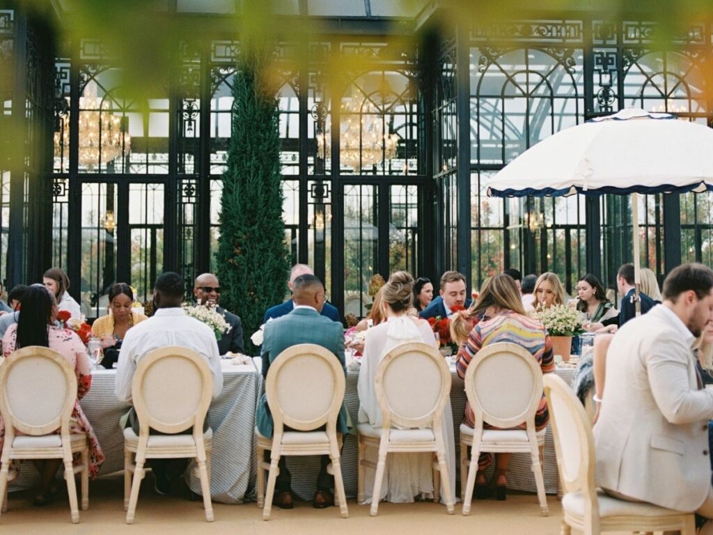Bride and groom seated at a table with wedding guests enjoying a meal in front of a glass building.